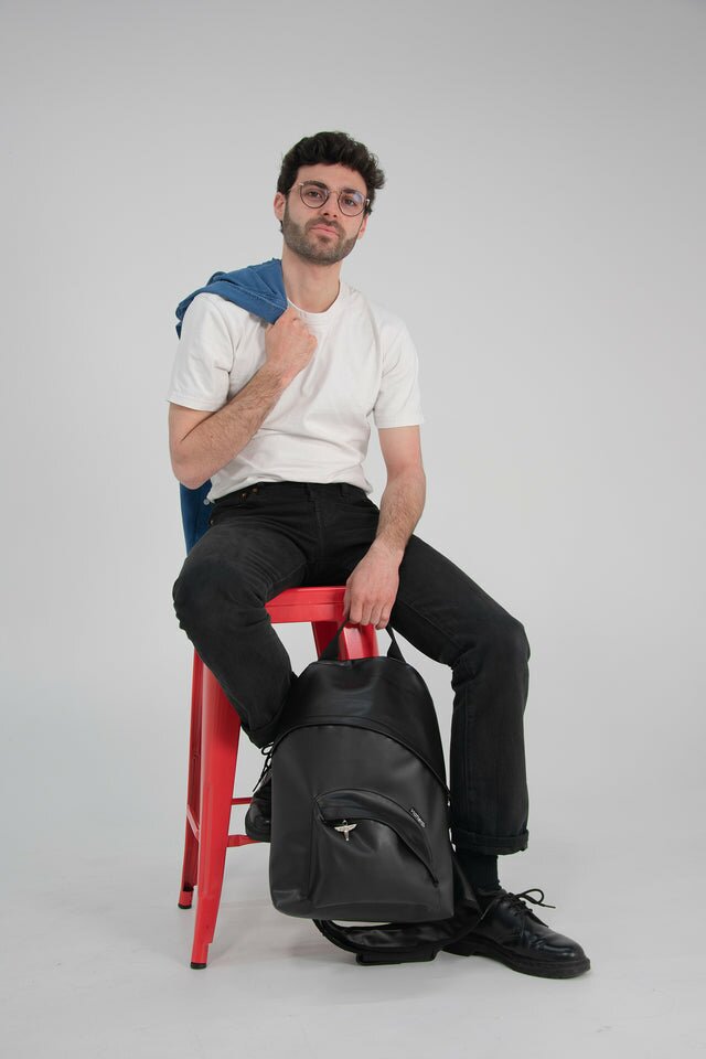 Photo mode homme streetwear. Un homme brun est assis sur un tabouret rouge industriel en metal. Il porte un tee-shirt blanc et un pantalon noir, l'un de ses pieds relevé sur l'un des rebords du tabouret. Il tient une chemise en jean bleu qui repose sur son épaule et dans son autre main, il tien la hanse du Pilot Bag. Sac à dos noir mono-bandoulière, made in france et vegan.